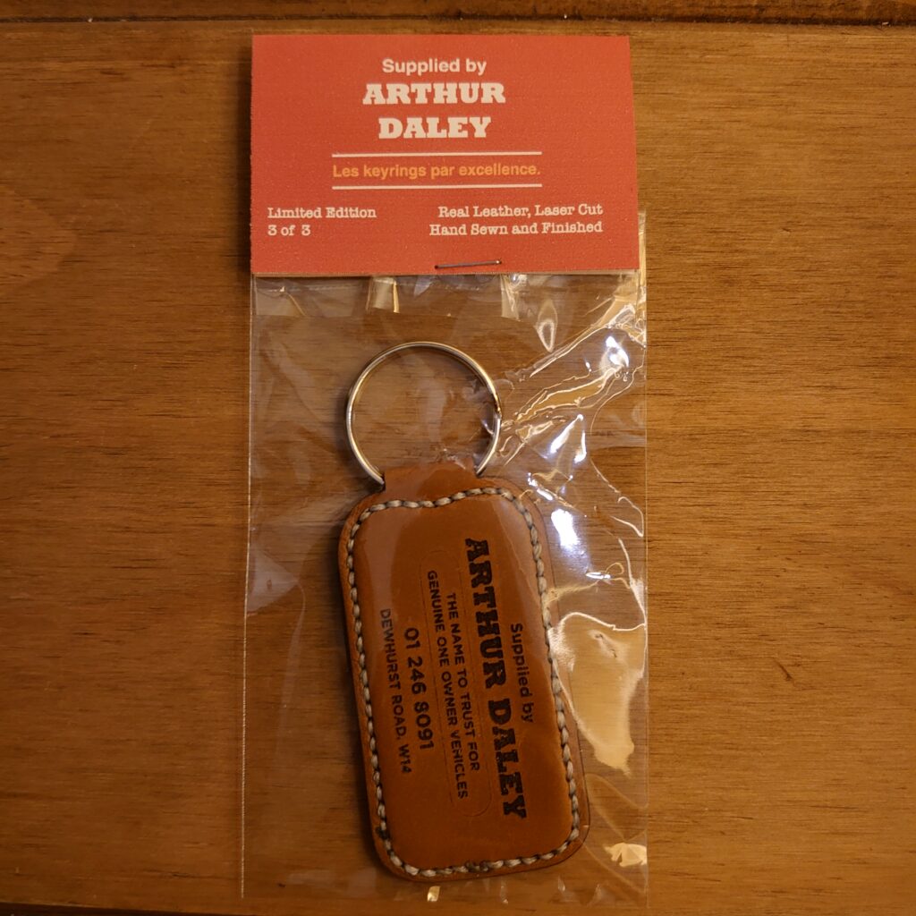 The keyring is adorned with the name "Arthur Daley" engraved in bold letters on the front, along with a laser-cut image of a vintage car. The leather is a rich brown color, and the edges are hand-sewn with white thread for a polished finish. The keyring is attached to a silver metal ring, making it easy to add to your keys or attach to a bag. It is presented in a retro-style bag, adding an extra touch of vintage charm to the product.
