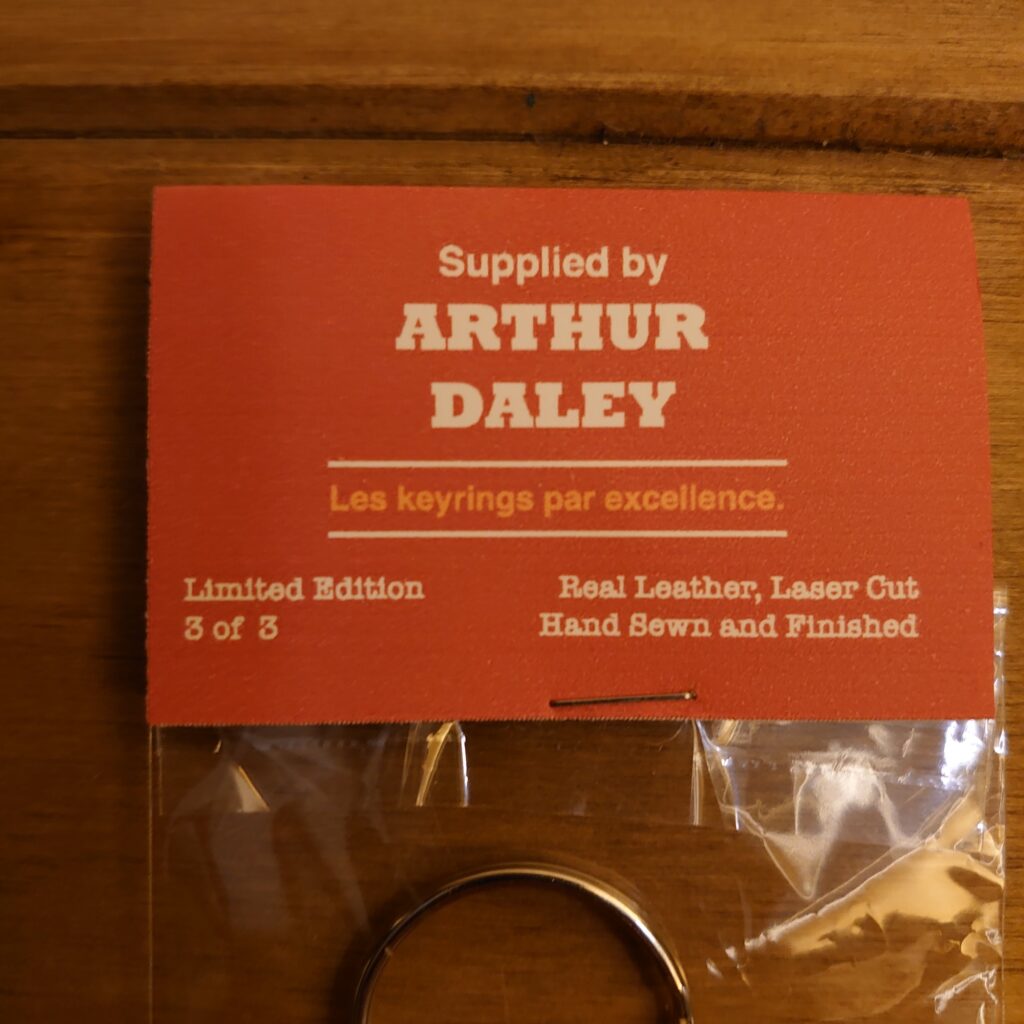 The keyring is adorned with the name "Arthur Daley" engraved in bold letters on the front, along with a laser-cut image of a vintage car. The leather is a rich brown color, and the edges are hand-sewn with white thread for a polished finish. The keyring is attached to a silver metal ring, making it easy to add to your keys or attach to a bag. It is presented in a retro-style bag, adding an extra touch of vintage charm to the product.